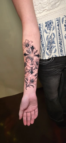 Fine line cherub hugging a cross with lilies, a forearm tattoo by Chyloe at Golden Tarot Ink Club