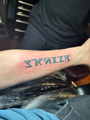 Bold stylized lettering across the forearm, a tattoo by Chyloe at Golden Tarot Ink Club