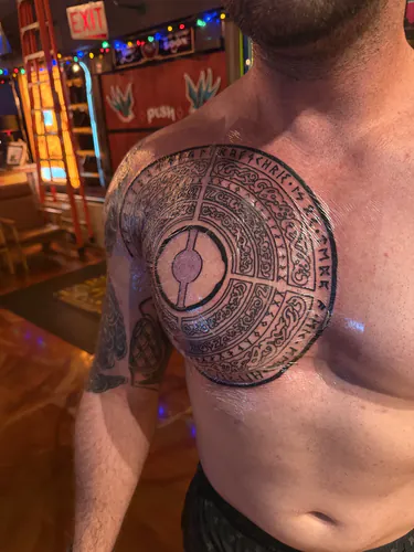 Large circular blackwork medallion tattoo ringed with stylized lettering across the chest and shoulder by Chyloe at Golden Tarot Ink Club